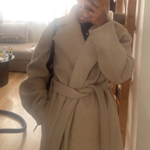 Beige kappa från NA-KD med bälte - Säljer en snygg beige kappa från NA-KD i storlek 36. Jackan har bred krage, knytbälte i midjan och dolda knappar framtill. Materialet är mjukt och känns lyxigt, perfekt för dig som gillar stilrena och klassiska plagg.