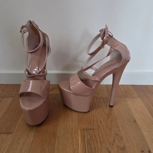 Rosa platåpumps med hög klack - Säljer ett par glansiga rosa pleasers med extremt hög klack och rejäl plattform. Skorna har öppen tå, smala remmar med spänne runt ankeln och dragkedja bak för enkel påtagning. Materialet är syntet med lackad finish som ger en snygg shine. Väldigt lätta i vikt vilket gör dem perfekta för dans. Tecken på användning finns.