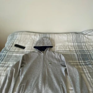Grå hoodie från Lacoste med dragkedja - Säljer en stilren grå hoodie från Lacoste med hel dragkedja och klassisk krokodillogga på bröstet. Tröjan har huva med mörkblå insida och två fickor framtill. Perfekt för en chill och sportig look.