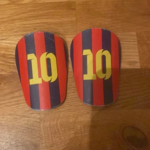 Shinpads med nummer 10, röd/blå - Ett par benskydd med röd och blå randigt mönster och siffran 10 i gult. Använda men i gott skick, med mindre repor och slitage. Perfekta för fotbollsträning eller match. Lätta och bekväma att bära.