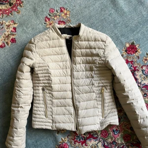 Beige pufferjacka från Rockandblue - Snygg beige pufferjacka från Rockandblue med guldfärgade dragkedjor och knappar. Jackan har quiltad design, rund krage och två fickor med dragkedja framtill. Perfekt för dig som vill ha en stilren och lätt jacka till kyligare dagar.