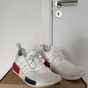 Adidas NMD vita sneakers med röd detalj - Adidas NMD sneakers i vitt med klassiska tre ränder på sidan, röda och blå detaljer på mellansulan och vit platt sula. Ovandelen är i syntetmaterial med snörning och rund tå. Skorna har en modern och sportig look, perfekt för dig som gillar streetwear.