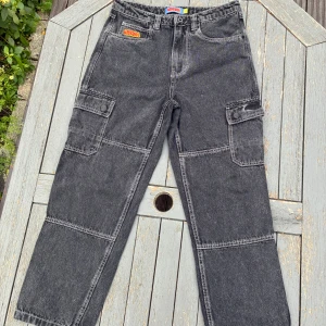 Svarta cargobyxor från Empyre - Svarta cargobyxor från Empyre med kontrastsömmar och broderad logga i rött och gult. Byxorna har flera stora fickor på sidorna och bak, raka ben och klassisk jeanslook. Perfekta för dig som gillar streetwear och praktiska detaljer.