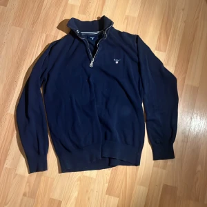 Marinblå half zip tröja från GANT - Snygg marinblå stickad tröja från GANT med halv dragkedja och broderad logga på bröstet. Tröjan har ribbade muddar, hög krage med randig detalj och lång ärm. Perfekt för dig som gillar klassisk och stilren look. Står 13-14/158-164, men är lite liten i storleken så mer 146/152. Följ mig så får ni 10kr rabatt!