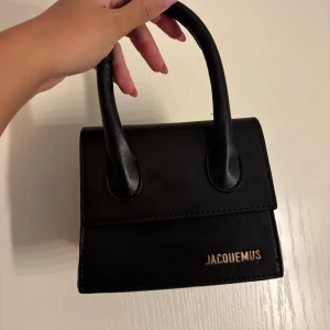 Ej äkta Svart Jacquemus handväska i skinn - Snygg svart handväska från Jacquemus med stilren, boxig form och två rejäla handtag. Väskan är tillverkad i slätt skinn och har en guldig Jacquemus-logga på framsidan. Perfekt storlek för det viktigaste och ger en cool vibe till din outfit. Använd men i fint skick
