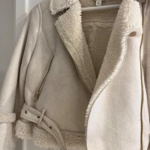Beige teddyjacka från Miss Selfridge - Supermjuk och snygg beige teddyjacka från Miss Selfridge med fuskpälsfoder, dragkedja, bälte i midjan och detaljer vid ärmslut och krage. Jackan har croppad passform och coola guldfärgade dragkedjor. Perfekt för kalla dagar när du vill vara både varm och trendig.