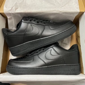 Nike Air Force 1 helsvarta sneakers - Säljer ett par klassiska Nike Air Force 1 i helsvart läder. Skorna har låg profil, rund tå, snörning och platt sula. Perfekta för dig som gillar en clean och tidlös look. Swoosh-loggan på sidorna och diskreta detaljer gör dem till ett måste i garderoben.