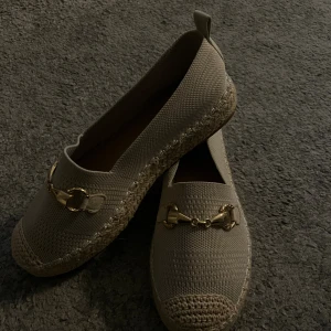 Beige loafers  - Snygga beige loafers med flätad sula och guldfärgad metallspänne framtill. Skorna har rund tå och är tillverkade i ett stickat syntetmaterial med grov textur. Perfekt för dig som gillar stilrena och trendiga skor med en twist.