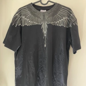 Svart t-shirt Marcelo Burlon med vingeprint - Svart t-shirt från Marcelo Burlon County of Milan med ikoniskt vingeprint i silvergrått över axlar och halsringning. Modellen är kortärmad och har en klassisk rund hals. Materialet är mjuk bomull som känns skön mot huden. Perfekt för dig som gillar streetwear med edge.