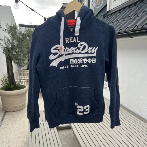 Mörkblå Superdry hoodie med tryck - Snygg mörkblå hoodie från Superdry med stort vitt tryck på bröstet och coola japanska tecken. Tröjan har huva med vita snören, känguruficka och ribbade muddar. Perfekt för dig som gillar streetwear och vill ha en chill look.