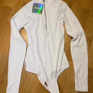 Vit ribbad bodysuit från Missguided - Säljer en vit ribbad bodysuit från Missguided med lång ärm och hög krage. Toppen har en snygg silverfärgad dragkedja framtill med ringdetalj. Perfekt att styla med jeans eller kjol för en clean look.