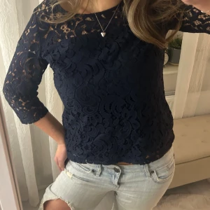 Marinblå spetstopp med trekvartsärm - Säljer en mörkblå topp i spets med snyggt mönstrad yta och trekvartsärmar. Toppen har rund halsringning. Säljer då jag har för mycket toppar som inte kommer till användning! 
