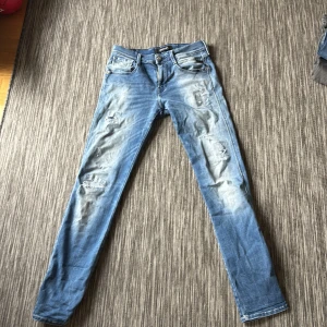 Replay Anbass Hyperflex jeans blå - Säljer ett par Replay Anbass Hyperflex jeans i blå tvätt med slitna detaljer och skinny passform. Byxorna har klassisk femficksdesign, knappgylf och stretchigt material som ger extra komfort. Perfekt för dig som gillar en smal siluett och coola slitningar.