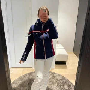 J.Lindeberg blå skidjacka med röda detaljer - Snygg blå skidjacka från J.Lindeberg med röda och vita detaljer, dragkedjor och flera fickor. Jackan har huva, lång ärm och sportig passform. Insidan är fodrad i vitt och svart med blå accenter och J.Lindeberg-logga. Perfekt för vinterns äventyr i backen. Perfekt skick, nypris 4600kr 