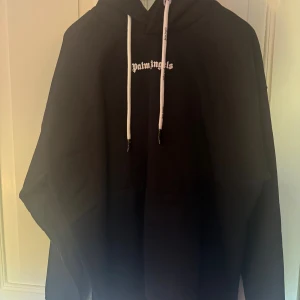Svart Palm Angels hoodie med tryck - Svart hoodie från Palm Angels med stor vit logotyp på ryggen och mindre tryck framtill. Tröjan har huva med vita snören.