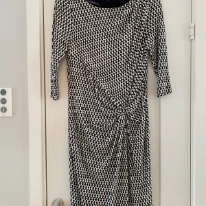 Svartvit mönstrad långklänning - Snygg långklänning från Lou&Joy med svartvitt rutigt mönster och cool knytning framtill i midjan. Klänningen har trekvartsärm och är gjord i mjukt, stretchigt material som sitter skönt. Perfekt för dig som gillar grafiska prints och vill sticka ut. Mått  Över bröstet: ca 47 cm  Över höften: ca 46 cm  Längd från axeln och ner: ca 106 cm   Klänningen: 100% viskos Underklänningen: 85% polyamid och 15% elastan   Kommer från ett djur och rökfritt hem. 