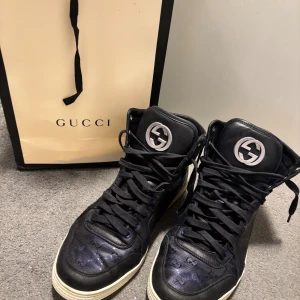 Gucci Ace High - Säljer ett par svarta high top sneakers från Gucci med GG-monogram i materialmix av skinn och textil. Skorna har snörning, vit sula och stora silverfärgade Gucci-loggor på plösen. Riktigt snygga och unika sneakers för dig som gillar lyxiga detaljer.Ovanlig variant av den höga sneakers från. Gucci. Färgen är svart på skinndetaljerna men blå-lila på sidorna.   Välbevarade. Innersulan saknas i övrigt mycket bra skick.   Storlek 7.5 us köpbevis från Sellpy finns. Båda påsar. Ingår