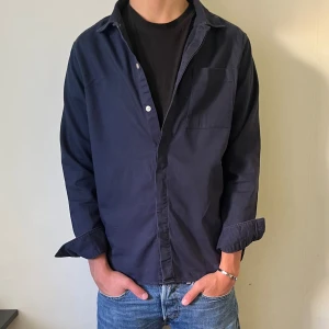 Mörkblå skjorta/overshirt från East West - Säljer en stilren mörkblå skjorta från East West. Skjortan/overshirt har klassisk krage, bröstficka och knappar framtill. Tillverkad i mjuk bomull och passar perfekt till jeans eller chinos. Enkel och clean look som funkar året runt.