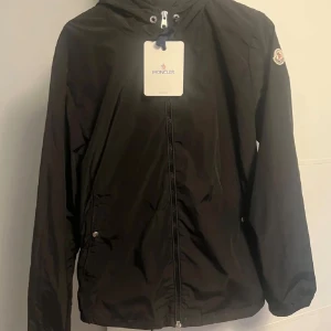 Svart vindjacka från Moncler - Svart vindjacka från Moncler med huva och dragkedja framtill. Jackan har Moncler-logga på ärmen, snörning vid huvan och diskreta fickor med tryckknapp. Tillverkad i lätt polyester, perfekt för blåsiga dagar.