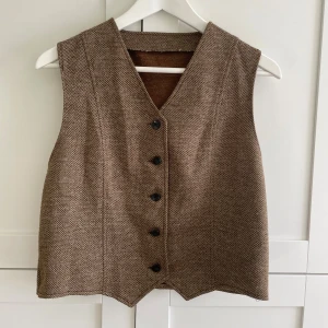 Brun väst / slipover  - Brun väst med v-ringning och svarta knappar framtill. Västen har en diskret rutig struktur och ett bälte baktill. Använt men bra skick - inga defekter. 