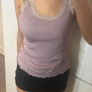 Ljuslila ribbad linnetopp med spets - Supersöt ljuslila ribbad linnetopp med breda spetskanter i beige runt halsringning och axlar. Toppen är ärmlös och har en figurnära passform med vågig kant nertill. Perfekt att styla med kjol eller jeans för en trendig look. Vet ej storlek men den är stretchig så passar nog de flesta!