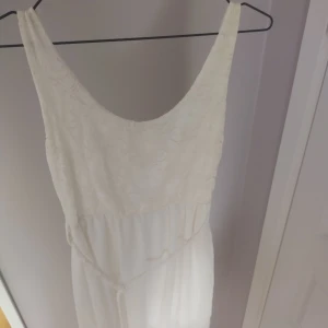 Vit långklänning med spetsdetaljer - Säljer en elegant vit långklänning med vacker spets upptill och ett flowy tyg nedtill. Klänningen har ärmlös design, rundad urringning och open back. Perfekt för sommarens speciella tillfällen. Spetsen har ett blommigt mönster som ger en romantisk känsla.