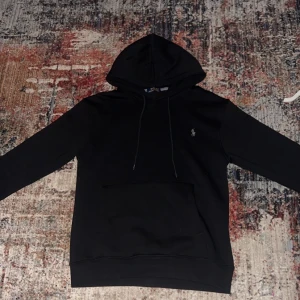 Svart hoodie från Polo Ralph Lauren - Svart hoodie från Polo Ralph Lauren med klassisk huva och snörning. Tröjan har en stor magficka framtill och den ikoniska Polo-loggan broderad på bröstet. Tillverkad i mjuk bomull för skön komfort och stilren look. Passar för M och S