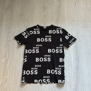 Svart BOSS t-shirt med logga, slim fit - Svart t-shirt från Hugo Boss i slim fit med vit BOSS-logga tryckt över hela plagget. Klassisk rund halsringning och korta ärmar. Tillverkad i mjuk bomull som känns skön mot huden. Perfekt för dig som gillar streetstyle och tydliga loggor.