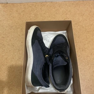 Mason Garmets sneakers i mocka - Snygga sneakers i svart och mörkblå mocka med vita sulor och svarta snören. Skorna har gulddetaljer vid snörningen och en klassisk rund tå. Perfekta för dig som gillar stilrena och bekväma skor med en modern touch.