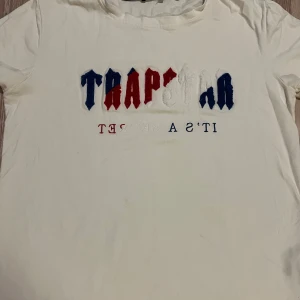 Vit Trapstar t-shirt med broderi - Vit t-shirt från Trapstar med broderad logga i blått, rött och vitt på bröstet. Texten 'IT'S A SECRET' är tryckt under loggan. Klassisk rund hals och korta ärmar. Perfekt för dig som gillar streetwear och vill sticka ut med coola detaljer. Passar även xs priset går att diskuteras 