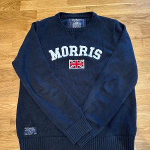 Mörkblå stickad tröja från Morris - Snygg mörkblå stickad tröja från Morris med broderad Union Jack-flagga och tydlig logga framtill. Tröjan har ribbade muddar vid ärmslut och nederkant samt rund hals. Väldigt fint skick! Skulle säga att den även passar om man har S som storlek. Kommer med originalkartong. 