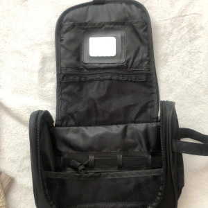 Svart necessär med dragkedja - Praktisk svart necessär med flera fack och dragkedja. Tillverkad i slitstark syntet och har smarta nätfickor för organisering. Perfekt att hänga upp tack vare ögla upptill. Enkel och stilren design.