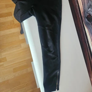 Svarta skinny byxor med dragkedja - Snygga svarta byxor i skinny modell med coola sömdetaljer och dragkedja vid benslutet. Materialet har en lätt glansig finish som ger en trendig look. Perfekta för dig som gillar en tight passform och stilrena detaljer.