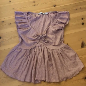 Rosa blus med volang från 157 - Supersöt ljuslila blus från 157 med volangdetaljer på axlarna och v-ringning. Blusen har knyt framtill och är tillverkad i ett mjukt, ribbat bomullsmaterial. Den har en flowy passform och korta ärmar som ger en chill vibe.