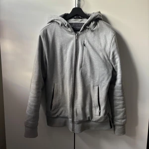 Grå jacka från Moose Knuckles - Snygg grå jacka från Moose Knuckles med svart logga på ryggen och metallogo på ärmen. Fodrad med quiltat svart innerfoder och gul detaljficka. Dragkedja framtill, två fickor och huva med snörning. Perfekt för kyliga dagar.