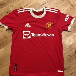Manchester United röd fotbollströja Adidas - Säljer en officiell Manchester United fotbollströja från Adidas i rött med vita detaljer. Tröjan har korta ärmar, rund hals och klubbmärke på bröstet. Materialet är lätt och andas, perfekt för träning eller match. TeamViewer-logga på framsidan och klassiska Adidas-ränder på axlarna.