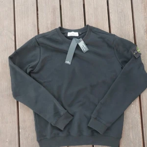 Svart sweatshirt från Stone Island - Svart sweatshirt från Stone Island med klassisk rund hals och ribbade muddar. Tröjan har den ikoniska Stone Island-loggan på vänster ärm och är tillverkad i mjuk bomull. Perfekt för dig som gillar streetwear och vill ha en clean look.