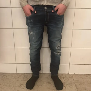Replay Anbass blå jeans med slitningar - Säljer ett par Replay Anbass jeans i mörkblå tvätt med snygga slitningar och trasiga detaljer på benen. Modellen är slim och har klassiska fem fickor med guldfärgade sömmar. Perfekt passform och cool look för dig som gillar streetstyle. W26 L30