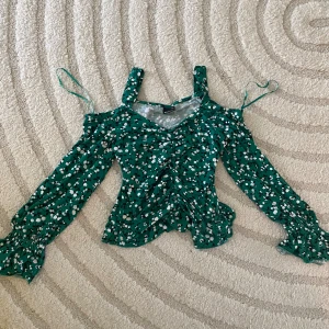 Grön blommig blus från Gina Tricot - Supersöt grön blus från Gina Tricot med vitt och svart blommönster. Toppen har v-ringning, långa ärmar med smock vid ärmslut och axelband. Snygg rynkdetalj framtill och lätt croppad passform. Perfekt för vår och sommar!