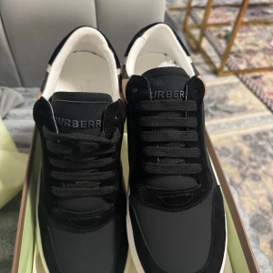Burberry rutig sneakers svart/vit 42 - Helt nya Snygga Burberry sneakers i svart med klassiskt rutigt mönster på sidorna i beige, svart och vit. Skorna har platt vit sula, rund tå och svarta snören. Materialmix av skinn och textil, med Burberry-logga på hälen och tungan. Perfekt för dig som gillar lyxiga detaljer och streetstyle.