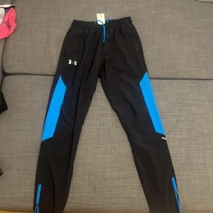 Svarta och blå träningsbyxor Under Armour - Snygga svarta träningsbyxor från Under Armour med blå detaljer längs benen och logga på låret. Byxorna har snörning i midjan och dragkedja vid benslut. Perfekta för sport eller chill, tillverkade i lätt polyester som andas.