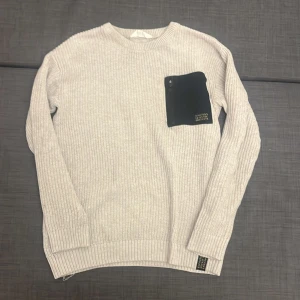 Beige stickad tröja med svart ficka H&M - Stickad beige tröja från H&M med långärmad passform och cool svart ficka med dragkedja på bröstet. Fickan har tryck i gult och ger en unik touch. Tröjan har ribbade muddar och är perfekt för dig som gillar stilrena detaljer.