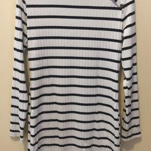 Randig ribbad långärmad tröja - Säljer en vit och svart ribbad långärmad tröja med horisontella ränder och snygga knappar på ena axeln. Tröjan har en klassisk passform och är perfekt för dig som gillar stilren och enkel design.