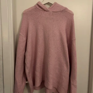 Ljusrosa stickad hoodie - Mysig ljusrosa stickad hoodie med huva och långa ärmar. Tröjan har en avslappnad passform och är perfekt för kyliga dagar. Ribbstickade muddar vid ärmslut och nederkant ger en extra touch. Enkel och stilren design som är lätt att matcha.