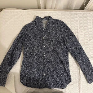 Blommig skjorta från Mango, slim fit - Mörkblå skjorta från Mango med vitt blommigt mönster och klassisk krage. Slim fit-modell med långa ärmar och vita knappar framtill. Perfekt för dig som gillar stilrena men ändå lekfulla detaljer.