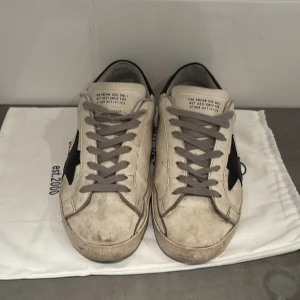 Golden Goose vita sneakers med svart stjärna - Golden Goose sneakers i vitt skinn med ikonisk svart stjärna på sidan och svart hälparti med logga. Grå snörning och beige detaljer vid tån. Platt sula och rund tå. Perfekt för dig som gillar en cool och avslappnad streetstil.