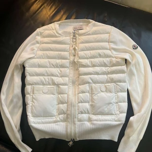 Moncler quiltad pufferjacka med dragkedja - Quiltad pufferjacka från Moncler med rund hals, två stora fickor framtill och dragkedja. Finns i både vit och svart, med Moncler-logga på ärmen. Jackan har stickade ärmar och ribbad nederkant för extra komfort. Perfekt för dig som gillar stilren och modern look.