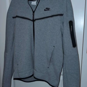 Grå ziptröja från Nike - Snygg grå ziptröja från Nike med svarta detaljer och dragkedja framtill. Tröjan har lång ärm, fickor med dragkedja och en extra ficka på ena ärmen. Perfekt för träning eller chill. Klassisk Nike-logga på bröstet.