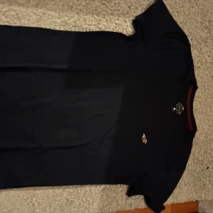 Mörkblå t-shirt från Hollister XS - Snygg mörkblå t-shirt från Hollister Must Have Collection i storlek XS. Klassisk rund hals med röd kant och diskret Hollister-logga på bröstet. T-shirten är kortärmad och gjord i mjuk bomull, perfekt för en chill och stilren look.