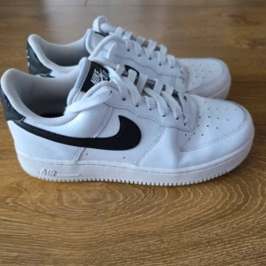 Nike Air Force 1 vita sneakers - Fräscha Nike Air Force 1 sneakers i vitt skinn med svart swoosh och klassisk siluett. Skorna har snörning, perforerad tå och låg profil. Perfekta för dig som vill ha en clean och tidlös look till vardags. Ikonisk streetstyle som aldrig går ur tiden. Perfekta inför höst och vinter då dem är väldigt varma och sköna.